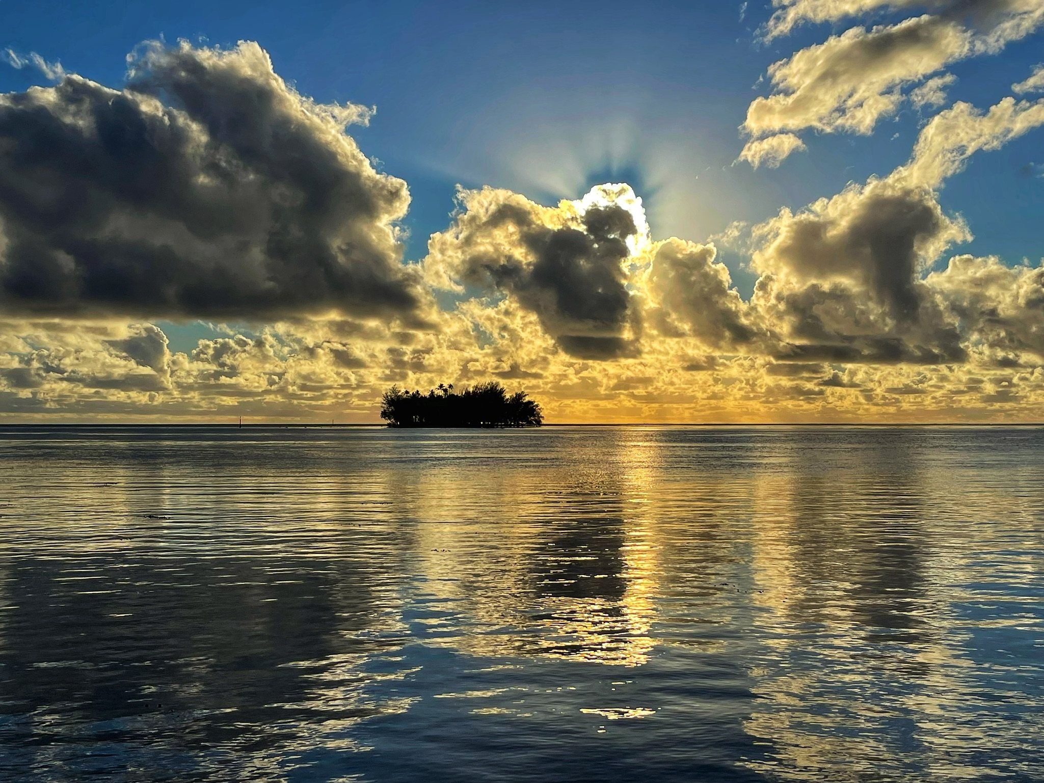 Sun breaking through banked clouds over a glassy lagoon, a small palm-covered islet on the horizon in French Polynesia.